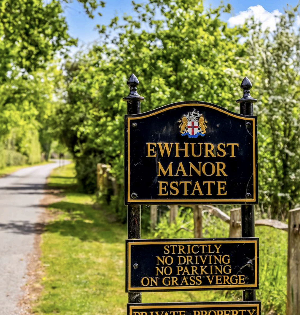 Black and gold Ewhurst Manor Estate sign with crest beside a hedged country lane