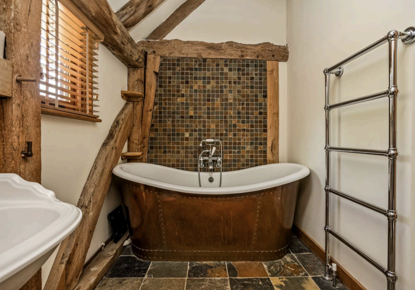 Bathroom with a copper roll top bath against a mosaic tiled wall, slate floor and chrome towel rail