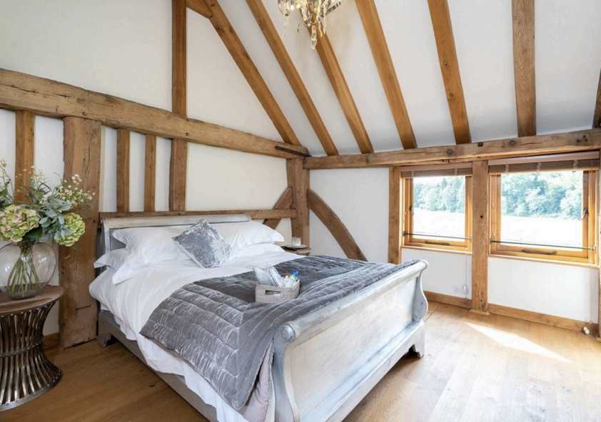 Bedroom with a silver grey wooden sleigh bed, grey velvet throw and windows beneath exposed rafters